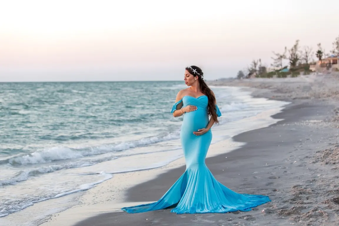 An expecting mom in a long blue dress on a quiet shoreline