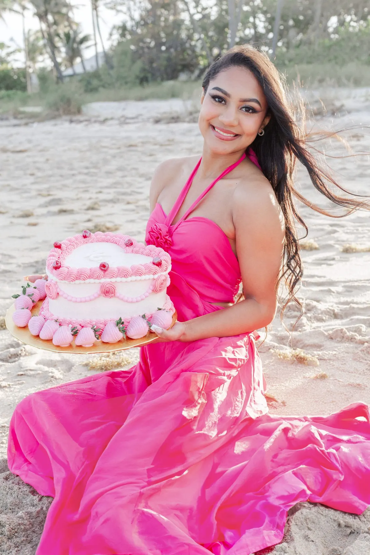 A woman in a pink dress holding a pink heart cake on the beach
